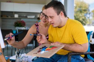 Teen boy with Down syndrome painting on canvas with his tutor    on terrace above riverbank